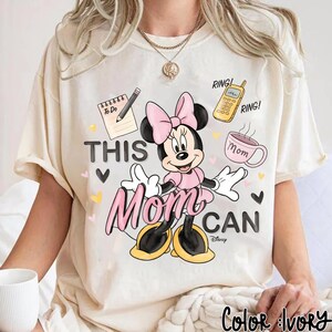 Może przedstawiać: Kremowa koszulka z Minnie Mouse w r&oacute;żowej sukience i kokardzie. Tekst "THIS Mom CAN" z ilustracjami notatnika, telefonu i filiżanki kawy. Widoczne logo Disneya.