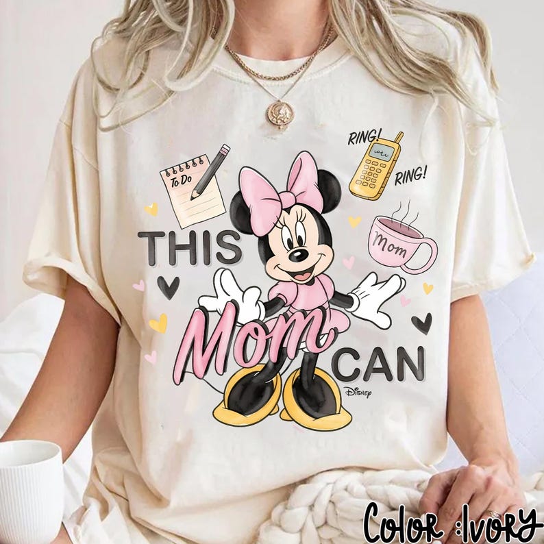 Może przedstawiać: Kremowa koszulka z Minnie Mouse w r&oacute;żowej sukience i kokardzie. Tekst "THIS Mom CAN" z ilustracjami notatnika, telefonu i filiżanki kawy. Widoczne logo Disneya.