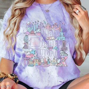 May include: A purple and white tie-dye t-shirt with a graphic of the Walt Disney World logo and park icons. The design includes Cinderella Castle, Spaceship Earth, and other attractions in pastel colours.