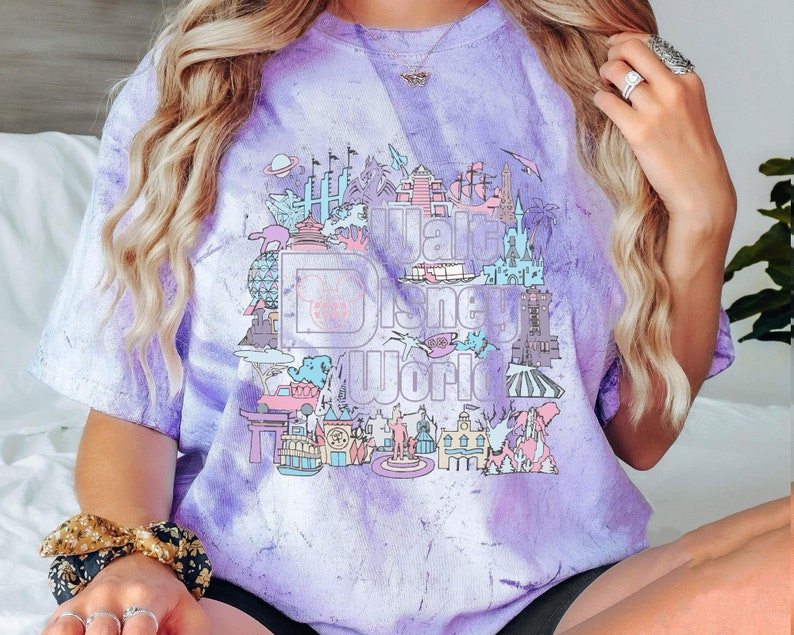 May include: A purple and white tie-dye t-shirt with a graphic of the Walt Disney World logo and park icons. The design includes Cinderella Castle, Spaceship Earth, and other attractions in pastel colours.