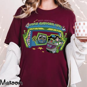 K&ouml;nnte beinhalten: Burgunderfarbenes T-Shirt mit einer farbenfrohen Grafik von Buzz Lightyears Space Ranger Spin. Das Design zeigt den Eingang der Attraktion, Buzz Lightyear und die kleinen gr&uuml;nen M&auml;nnchen. Das Shirt hat auch den Text "Buzz Lightyear's Space Ranger Spin".