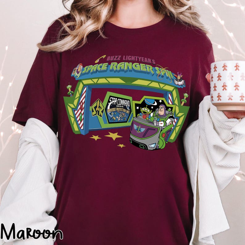 K&ouml;nnte beinhalten: Burgunderfarbenes T-Shirt mit einer farbenfrohen Grafik von Buzz Lightyears Space Ranger Spin. Das Design zeigt den Eingang der Attraktion, Buzz Lightyear und die kleinen gr&uuml;nen M&auml;nnchen. Das Shirt hat auch den Text "Buzz Lightyear's Space Ranger Spin".