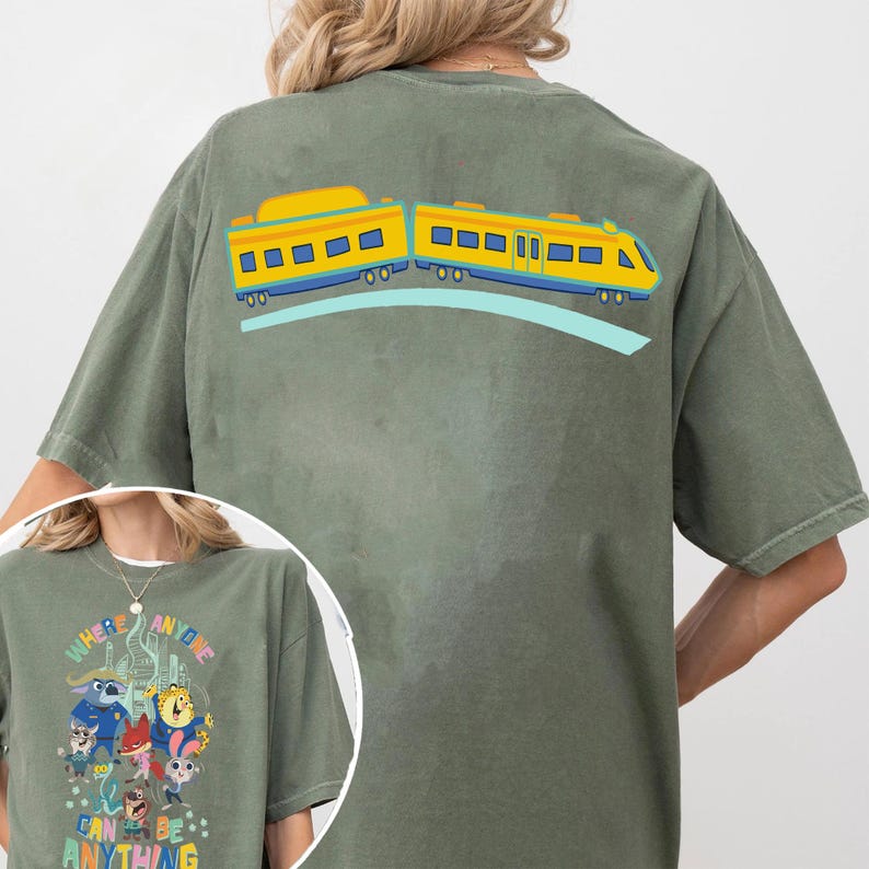 Pu&ograve; includere: T-shirt verde oliva con un design di treno giallo e blu sul retro, su un binario azzurro. La parte anteriore della maglietta presenta una grafica colorata con la scritta "WHERE ANYONE CAN BE ANYTHING".