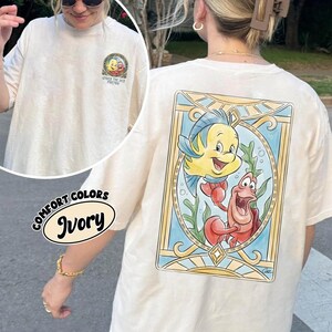 May include: Ivory-colored t-shirt featuring a colorful graphic of Flounder and Sebastian from The Little Mermaid. The shirt has a crew neck and short sleeves. The text "UNDER THE SEA FOREVER" is printed on the front.