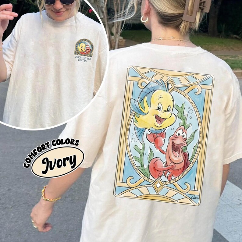May include: Ivory-colored t-shirt featuring a colorful graphic of Flounder and Sebastian from The Little Mermaid. The shirt has a crew neck and short sleeves. The text "UNDER THE SEA FOREVER" is printed on the front.