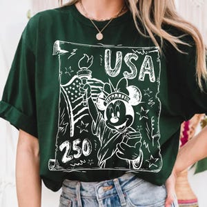 May include: Dark green t-shirt featuring a white graphic of Minnie Mouse as the Statue of Liberty, holding a torch and a book, with the text "USA" and "250". The design includes the American flag and stars.