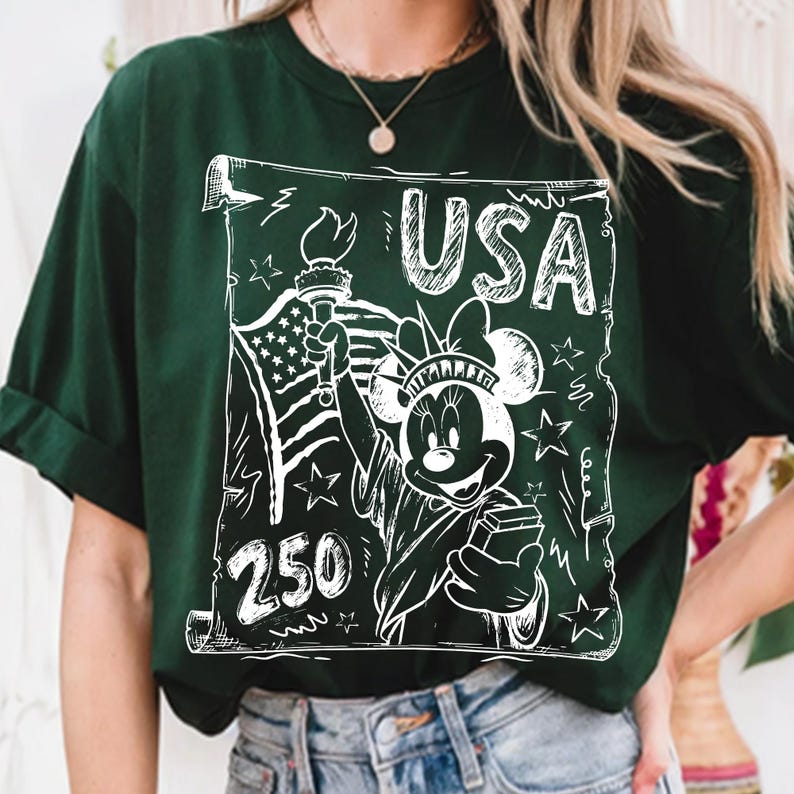 May include: Dark green t-shirt featuring a white graphic of Minnie Mouse as the Statue of Liberty, holding a torch and a book, with the text "USA" and "250". The design includes the American flag and stars.