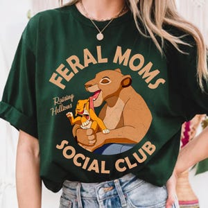 May include: Dark green t-shirt with the text "FERAL MOMS SOCIAL CLUB" in a circular design. The shirt features a cartoon lioness and cub. The words "Raising Hellions" are also printed on the shirt.
