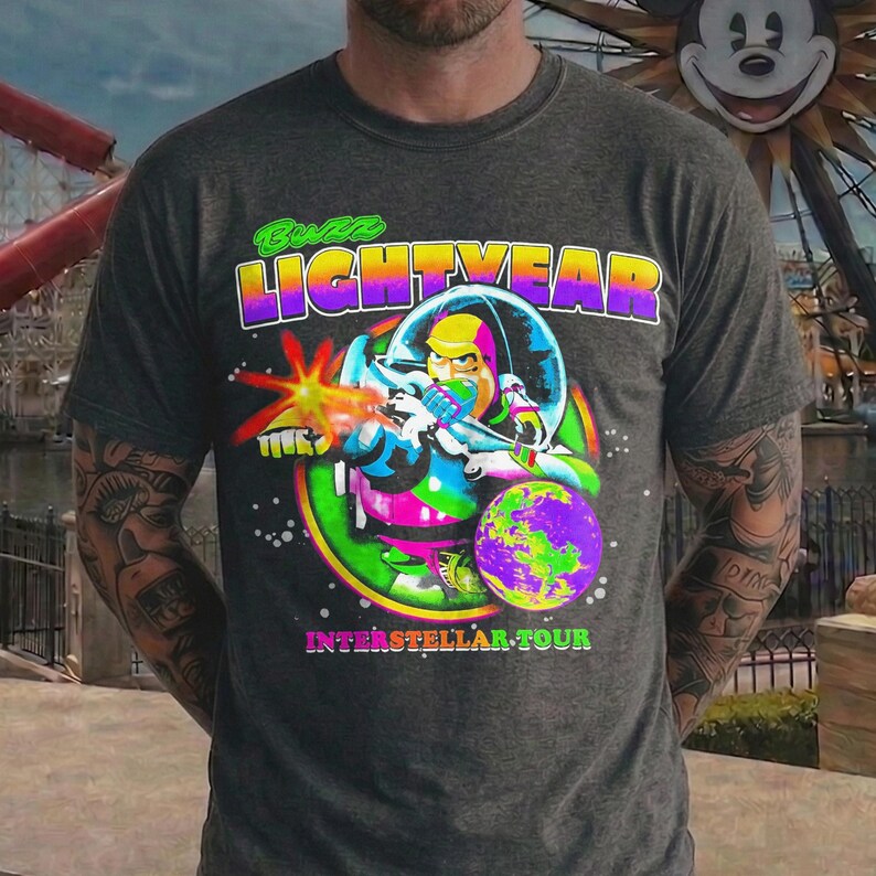 May include: Dark gray t-shirt featuring a colorful graphic of Buzz Lightyear in a space suit, firing a laser. The text "Buzz Lightyear" is above the image, and "Interstellar Tour" is below. The design includes a planet and a neon green and pink border.