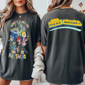 Pu&ograve; includere: T-shirt grigie scure con una grafica colorata. La parte anteriore della maglietta dice "WHERE ANYONE CAN BE ANYTHING" con personaggi dei cartoni animati ed edifici cittadini. Il retro presenta un treno giallo e blu.