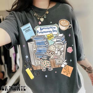 Puede incluir: Camiseta gris grafito con una ilustraci&oacute;n caprichosa para el D&iacute;a de los Profesionales Administrativos 2026. El dise&ntilde;o incluye un conejo de dibujos animados, pilas de papeleo y el texto "Assistant Mayor."