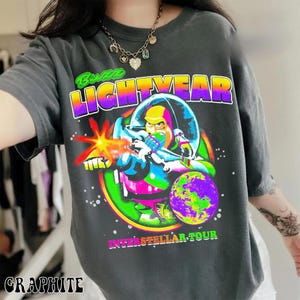 May include: Graphite-colored t-shirt featuring a vibrant graphic of Buzz Lightyear in a space suit, firing a laser. The design includes the text "Buzz Lightyear" in a colorful gradient, with "Interstellar Tour" below. The shirt is a crew neck.