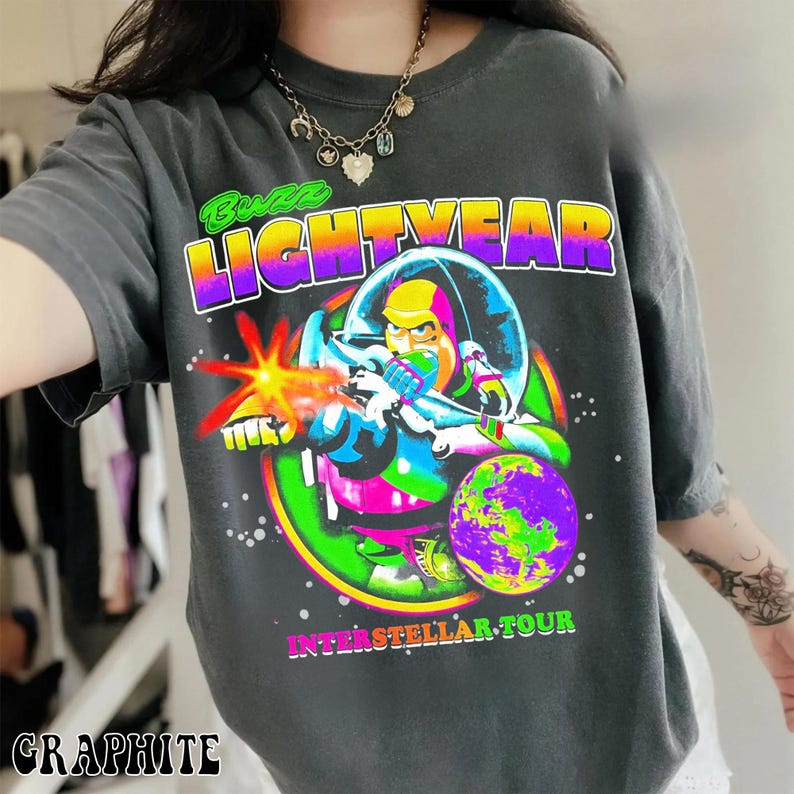 May include: Graphite-colored t-shirt featuring a vibrant graphic of Buzz Lightyear in a space suit, firing a laser. The design includes the text "Buzz Lightyear" in a colorful gradient, with "Interstellar Tour" below. The shirt is a crew neck.