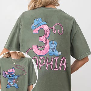 May include: Olive green t-shirt featuring a pink number 3 with a paisley design, a cowboy hat, and a blue boot with a cartoon character. The name "Sophia" is printed below the number in pink. A smaller image of the character is in the corner.