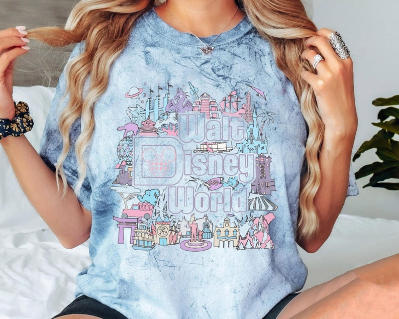 May include: A light blue tie-dye t-shirt with a colourful graphic of the Walt Disney World logo and park icons. The design includes the park's name, Cinderella's castle, and other attractions. The shirt has short sleeves.