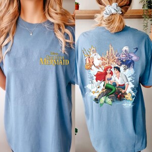 May include: A light blue t-shirt with a graphic print of characters from the Disney movie "The Little Mermaid". The front of the shirt has the movie title in yellow lettering.