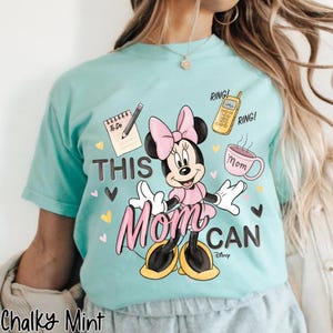 Może przedstawiać: Jasnoniebieska koszulka z grafiką Myszki Minnie w kolorze r&oacute;żowym i czarnym. Tekst "THIS MOM CAN" jest wyświetlany z ilustracjami listy rzeczy do zrobienia, telefonu i filiżanki kawy. Koszulka jest oznaczona jako "Chalky Mint".