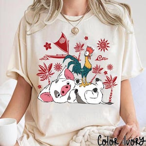May include: Ivory-colored t-shirt featuring a cartoon illustration of a pig and a rooster. The pig is white with pink ears and snout, and the rooster has blue and green feathers. The shirt also has red and white decorative elements.