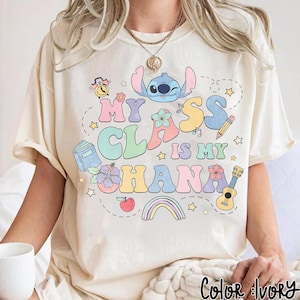 Puede incluir: Camiseta de color crema con un dise&ntilde;o gr&aacute;fico colorido. El dise&ntilde;o presenta el texto "My Class is My Ohana" con un personaje de dibujos animados, flores, un arco&iacute;ris y elementos escolares. La camiseta tiene un corte relajado.
