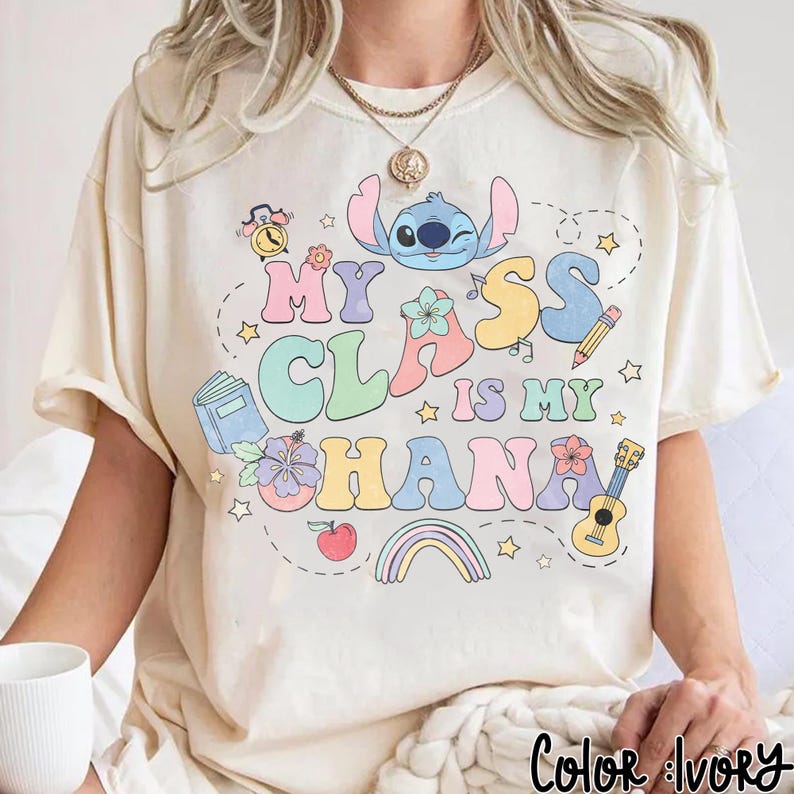 Puede incluir: Camiseta de color crema con un dise&ntilde;o gr&aacute;fico colorido. El dise&ntilde;o presenta el texto "My Class is My Ohana" con un personaje de dibujos animados, flores, un arco&iacute;ris y elementos escolares. La camiseta tiene un corte relajado.