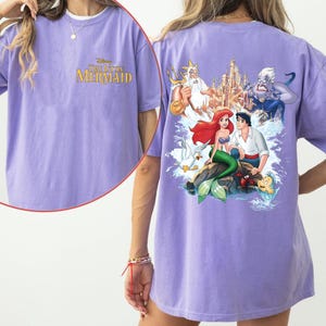 May include: Purple t-shirt with a graphic print of characters from the Disney movie The Little Mermaid. The text "Disney The Little Mermaid" is printed on the front of the shirt.