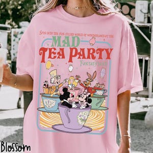 Puede incluir: Camiseta rosa claro con un gr&aacute;fico colorido de la Fiesta del T&eacute; del Sombrerero Loco de Wonderland. El dise&ntilde;o incluye a Alicia, el Sombrerero Loco y Mickey y Minnie Mouse en tazas de t&eacute;. El texto dice "Mad Tea Party Fantasy Land".