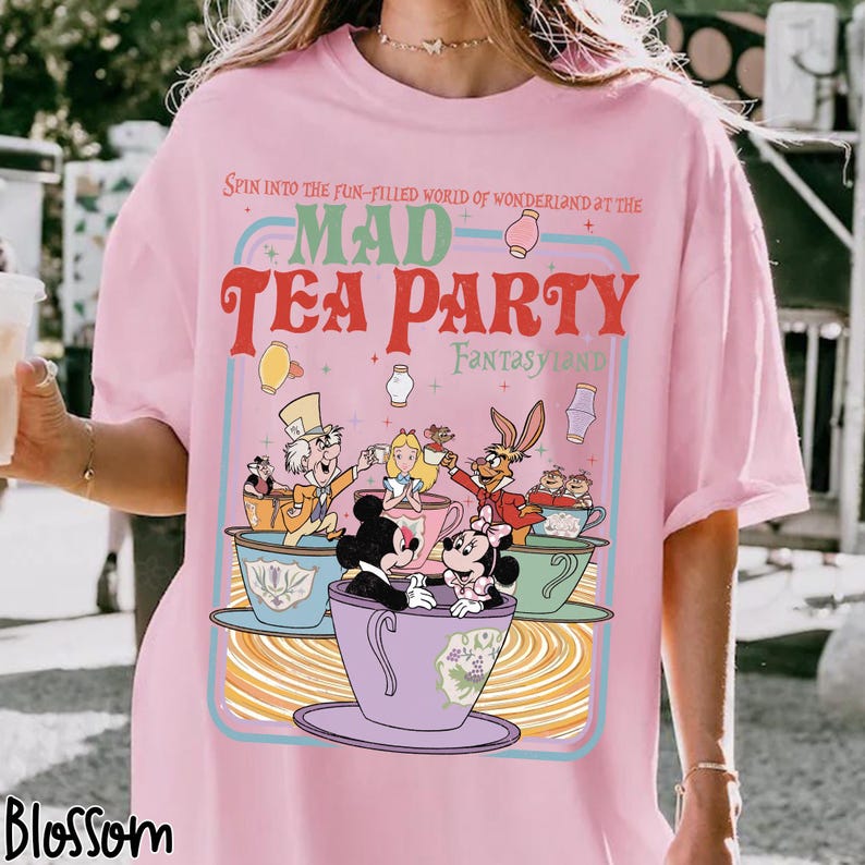 Puede incluir: Camiseta rosa claro con un gr&aacute;fico colorido de la Fiesta del T&eacute; del Sombrerero Loco de Wonderland. El dise&ntilde;o incluye a Alicia, el Sombrerero Loco y Mickey y Minnie Mouse en tazas de t&eacute;. El texto dice "Mad Tea Party Fantasy Land".