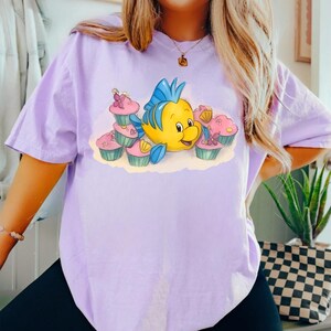 May include: A light purple t-shirt featuring a cartoon illustration of a yellow fish with blue fins and a smiling face, surrounded by pink cupcakes. The cupcakes have pink frosting and are decorated with flowers. The shirt is a casual style.