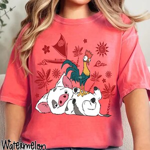 May include: A coral-colored t-shirt featuring a graphic of a pig and a rooster, with a sailboat and tropical elements. The word "Watermelon" is printed at the bottom left. The design is in shades of white, black, red, yellow, and blue.