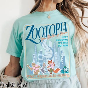 Puede incluir: Camiseta verde menta con un dise&ntilde;o de la pel&iacute;cula Zootopia. El dise&ntilde;o incluye la palabra "Zootopia" en azul, junto con las frases "You Belong Here", "Stay Pawsitive" y "It's Wild Out Here". La camiseta tambi&eacute;n tiene personajes de dibujos animados.