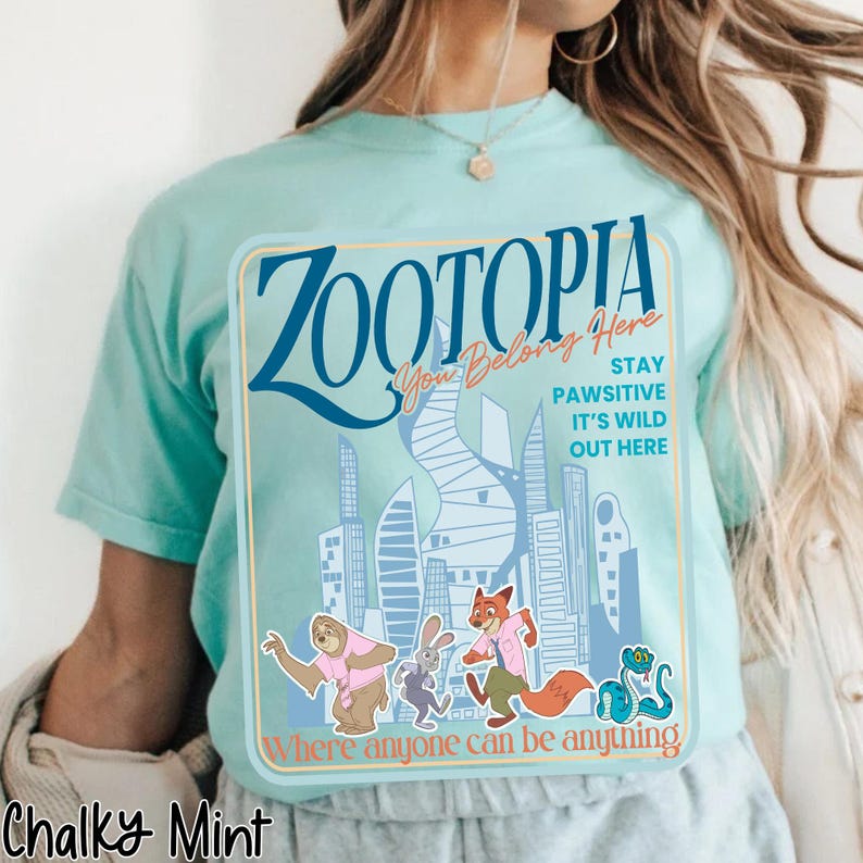 Puede incluir: Camiseta verde menta con un dise&ntilde;o de la pel&iacute;cula Zootopia. El dise&ntilde;o incluye la palabra "Zootopia" en azul, junto con las frases "You Belong Here", "Stay Pawsitive" y "It's Wild Out Here". La camiseta tambi&eacute;n tiene personajes de dibujos animados.