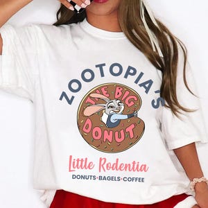 Puede incluir: Camiseta blanca con una imagen de dibujos animados de Judy Hopps de Zootopia dentro de una rosquilla. El texto de la camiseta dice "Zootopia's The Big Donut" y "Little Rodentia" con "Donuts, Bagels, Coffee" debajo.