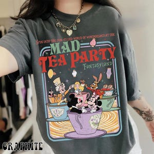 Puede incluir: Camiseta de color grafito con un dise&ntilde;o colorido de la Fiesta del T&eacute; del Sombrerero Loco de Wonderland. El dise&ntilde;o incluye personajes como el Sombrerero Loco, Alicia y la Liebre de Marzo en tazas de t&eacute;. El texto dice "MAD TEA PARTY FantasyLAND".