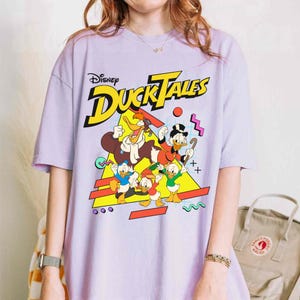 May include: A purple t-shirt with a graphic print of the Disney DuckTales characters. The text "Disney DuckTales" is printed in yellow and the characters are in a retro 90s style.