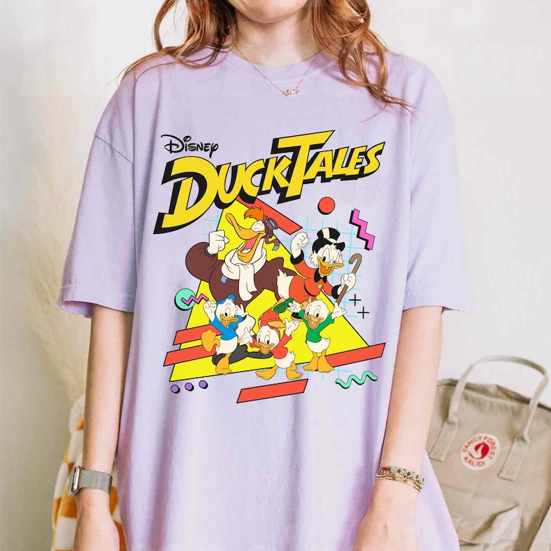 May include: A purple t-shirt with a graphic print of the Disney DuckTales characters. The text "Disney DuckTales" is printed in yellow and the characters are in a retro 90s style.