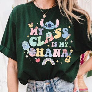 Puede incluir: Camiseta verde oscuro con la frase "My Class is My Ohana" en letras coloridas. El dise&ntilde;o incluye un personaje de dibujos animados, flores, un libro, un l&aacute;piz, una guitarra, un arco&iacute;ris y una manzana. La camiseta es de cuello redondo.