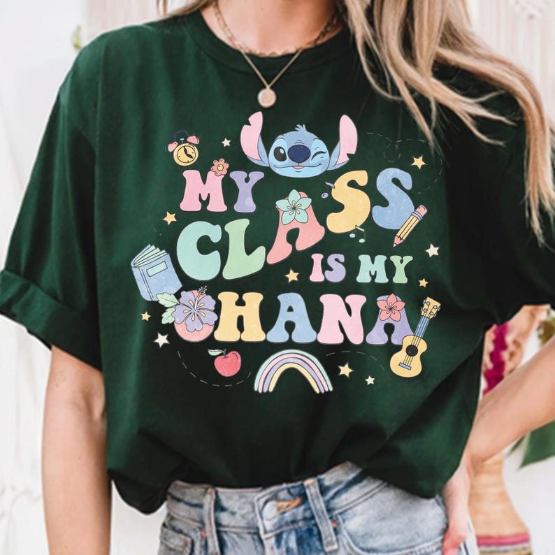 Puede incluir: Camiseta verde oscuro con la frase "My Class is My Ohana" en letras coloridas. El dise&ntilde;o incluye un personaje de dibujos animados, flores, un libro, un l&aacute;piz, una guitarra, un arco&iacute;ris y una manzana. La camiseta es de cuello redondo.