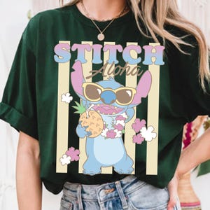 K&ouml;nnte beinhalten: Dunkelgr&uuml;nes T-Shirt mit einer Cartoon-Figur Stitch, die eine Sonnenbrille tr&auml;gt und eine Ananas h&auml;lt. Der Text "STITCH Aloha" wird &uuml;ber der Figur in einer bunten Schrift angezeigt, mit vertikalen Streifen im Hintergrund.