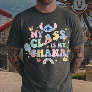 Puede incluir: Camiseta gris oscuro con la frase "My Class is My Ohana" en letras coloridas y divertidas. El dise&ntilde;o incluye un personaje de dibujos animados de Stitch, flores, un arco&iacute;ris y gr&aacute;ficos tem&aacute;ticos escolares. La camiseta es de cuello redondo.