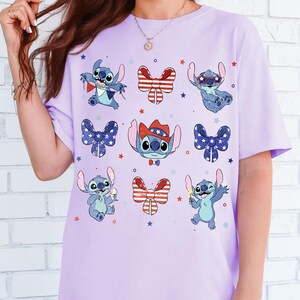 May include: A lilac t-shirt with cartoon Stitch characters in patriotic designs. Stitch is shown with sunglasses, holding a sparkler, and wearing a cowboy hat. Red, white, and blue bow ties and stars are also featured.