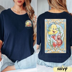 May include: Navy blue t-shirt featuring a colorful graphic of characters from "The Little Mermaid". The front has a small image with text, while the back displays a larger, detailed illustration of Flounder and Sebastian. The shirt is a comfortable, casual style.