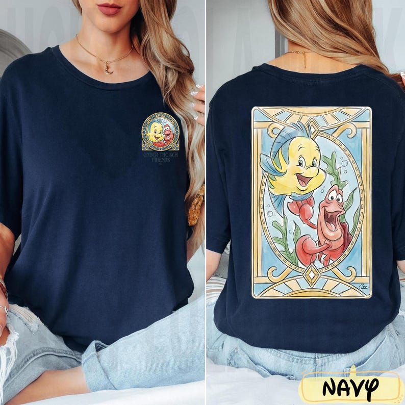 May include: Navy blue t-shirt featuring a colorful graphic of characters from "The Little Mermaid". The front has a small image with text, while the back displays a larger, detailed illustration of Flounder and Sebastian. The shirt is a comfortable, casual style.