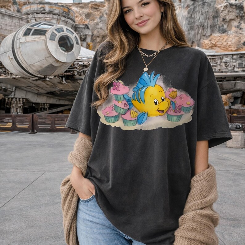 May include: Dark gray t-shirt featuring a cartoon image of a yellow fish with blue fins and a smiling face, surrounded by pink cupcakes. The shirt has a relaxed fit and short sleeves.