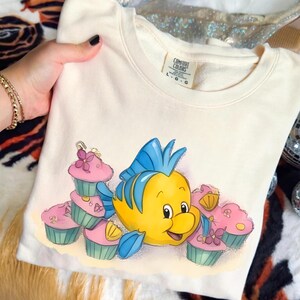 May include: Cream-colored sweatshirt featuring a cartoon illustration of a yellow fish with blue fins and a smiling expression, surrounded by pink cupcakes with blue and green accents. The sweatshirt has a Comfort Colors tag.