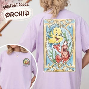 May include: Orchid-colored t-shirt featuring a colorful illustration of Flounder and Sebastian from The Little Mermaid. The back of the shirt showcases the characters within a stained-glass style frame. The front of the shirt has a smaller graphic with the text "Under the Sea Friends".