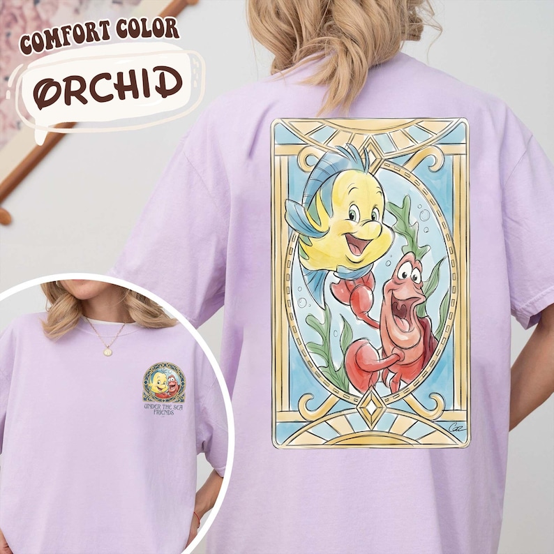 May include: Orchid-colored t-shirt featuring a colorful illustration of Flounder and Sebastian from The Little Mermaid. The back of the shirt showcases the characters within a stained-glass style frame. The front of the shirt has a smaller graphic with the text "Under the Sea Friends".