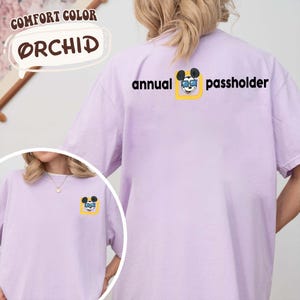 Puede incluir: Una camiseta de color lila claro con el texto "annual passholder" y un gr&aacute;fico de Mickey Mouse. La camiseta tambi&eacute;n tiene las palabras "Comfort Color" y "Orchid".