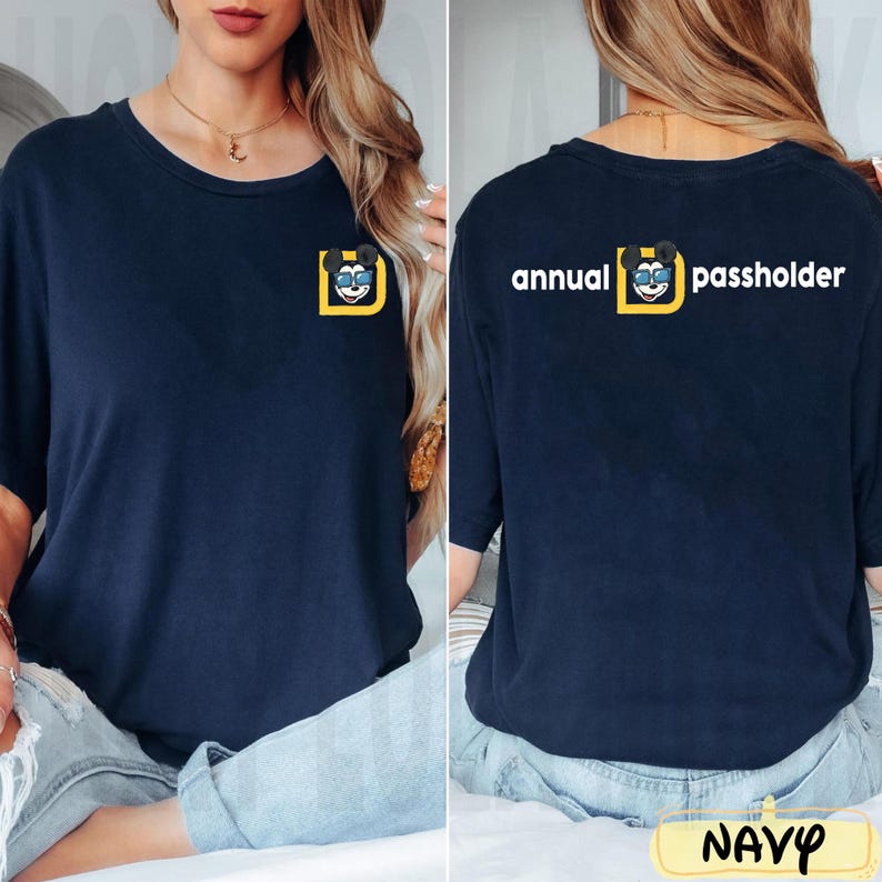 Puede incluir: Camiseta azul marino con un peque&ntilde;o gr&aacute;fico de un personaje de dibujos animados con gafas de sol dentro de un cuadrado amarillo. La parte posterior de la camiseta muestra el texto "annual passholder" encima del mismo gr&aacute;fico.