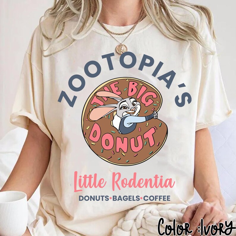 Puede incluir: Camiseta color marfil con un dibujo animado de Judy Hopps de Zootopia dentro de una rosquilla. El texto "ZOOTOPIA'S THE BIG DONUT" est&aacute; encima de la rosquilla, con "Little Rodentia" y "DONUTS &bull; BAGELS &bull; COFFEE" debajo. La camiseta es de corte holgado.