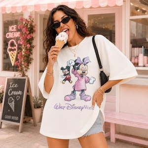 May include: A white t-shirt featuring a graphic of Minnie Mouse and Mickey Mouse with the text "Walt Disney World". The shirt is oversized and has short sleeves. The person in the image is wearing sunglasses and eating an ice cream cone.